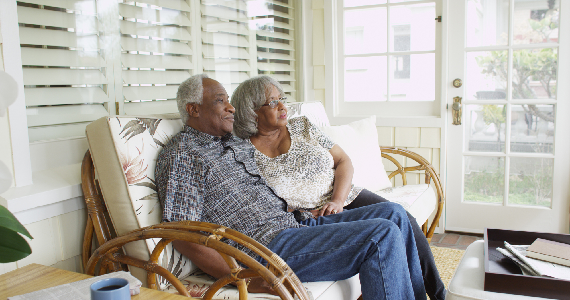 An elderly black couple sit on a rocker in a sunroom.